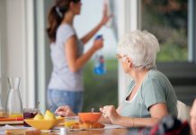 Senior Woman Having Breakfast With Home Care In The Background