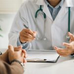 A professional physician in a white medical uniform talks to discuss results or symptoms and gives a recommendation to a male patient and signs a medical paper at an appointment visit in the clinic.
