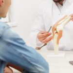 Doctor showing flexible knee joint model to his patient and explains the cause of his pain in medical office. Patient visiting physiotherapist in clinic. Surgery, healthcare and medicine concept